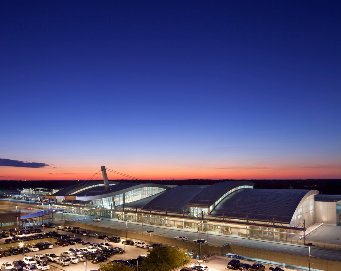 A modern airport terminal with curved roofs is illuminated at dusk, surrounded by parked cars and a glowing sky transitioning from orange near the horizon to deep blue above.