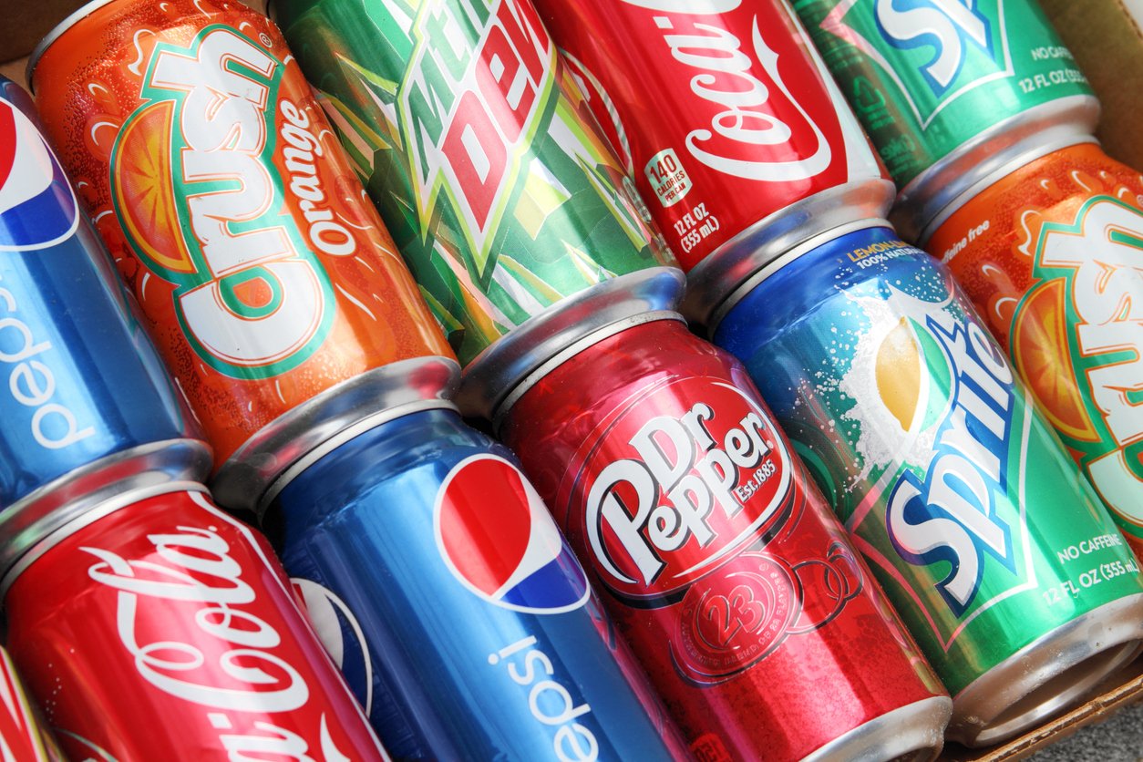 A close-up of assorted soda cans, including Coca-Cola, Pepsi, Dr Pepper, Mountain Dew, Crush Orange, and Sprite, arranged in rows in a cardboard box.