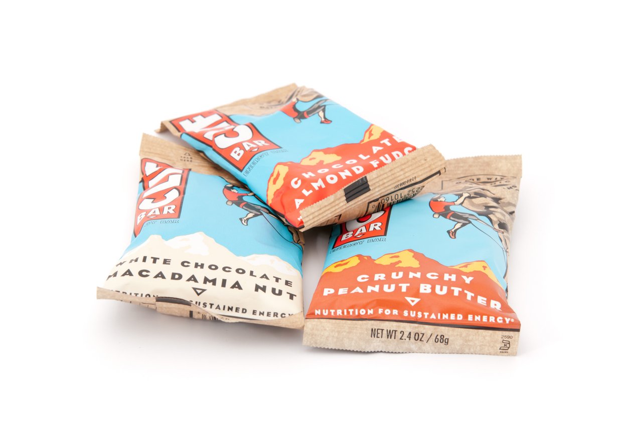 Three Clif Bars in their wrappers are stacked on a white background. The visible flavors are White Chocolate Macadamia Nut, Crunchy Peanut Butter, and Chocolate Almond Fudge.