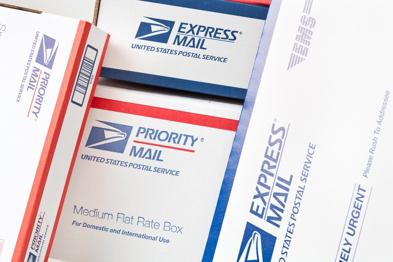 A close-up of several stacked USPS shipping boxes, including Priority Mail and Express Mail options, showing their labels and logos.