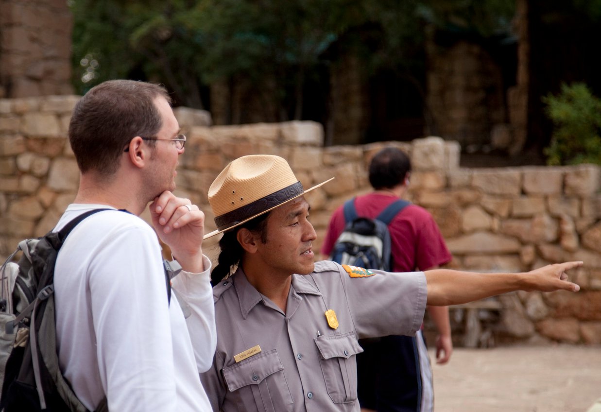 A park ranger in uniform and hat points while talking to a man with a backpack and glasses; they stand outdoors near a stone wall with trees in the background.
