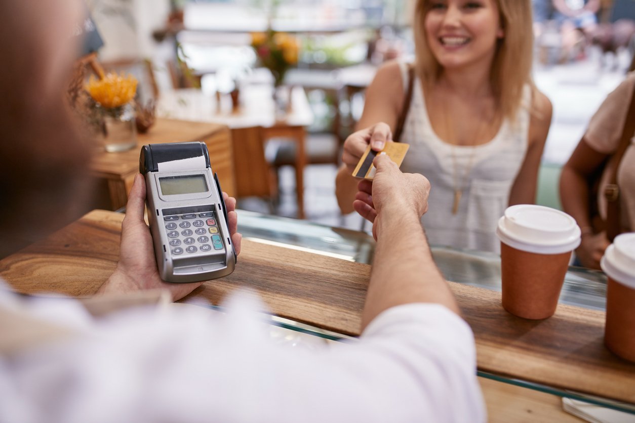 A customer smiles while handing a credit card to a cashier holding a payment terminal across a café counter. A takeaway coffee cup sits on the counter in front of her.