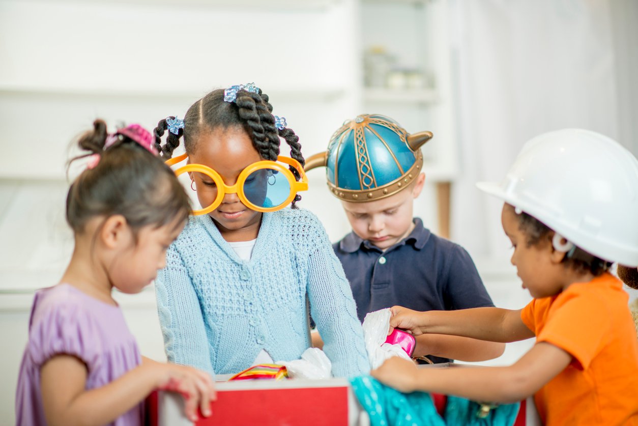 Four young children play dress-up. One girl wears large yellow glasses, another wears a white hard hat, one has a Viking helmet, and the fourth child is looking at costumes in a box. They are in a bright indoor setting.