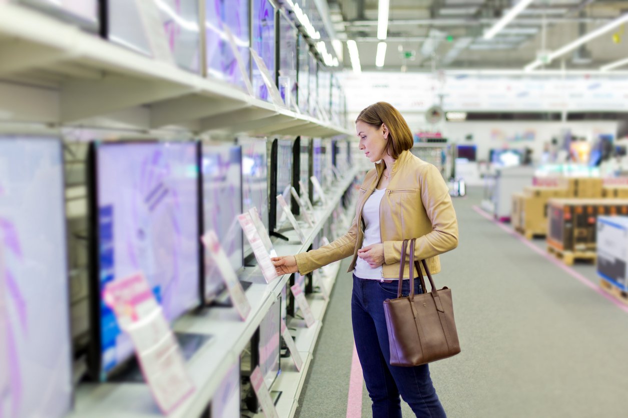 A woman in a tan jacket and jeans stands in an electronics store, holding a brown handbag and looking at information tags on flat-screen TVs displayed on shelves.