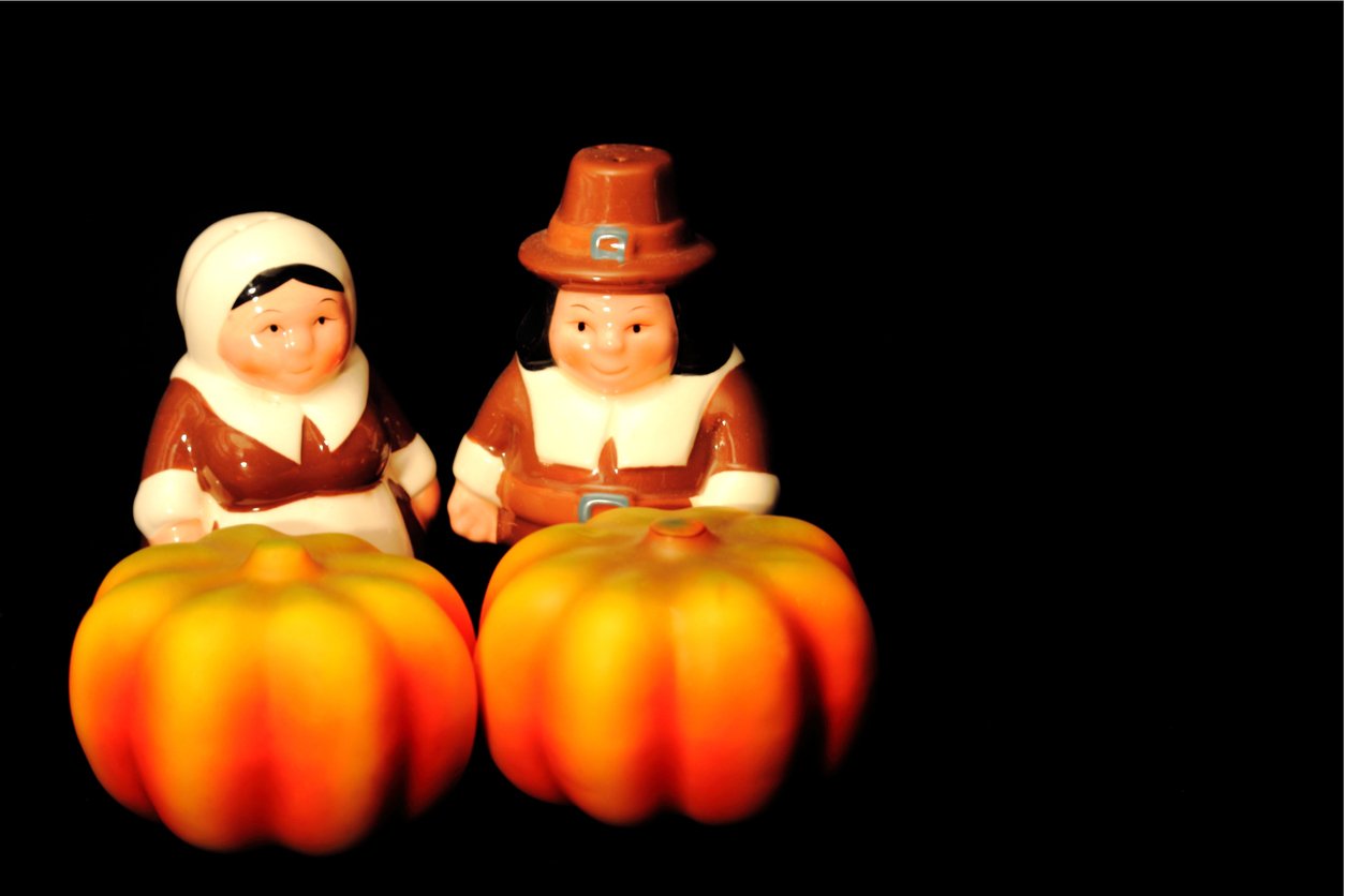 Two ceramic figurines dressed as Pilgrims, one male and one female, stand side by side behind two small decorative pumpkins against a black background, evoking the tradition and nostalgia of Thanksgiving Dinner Cost History.