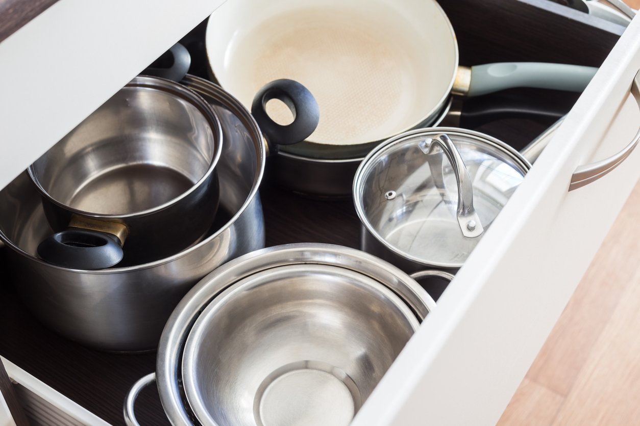 Open kitchen drawer containing stainless steel pots, a glass lid, and a frying pan with a light-colored interior and green handle. The items are neatly arranged inside the drawer.