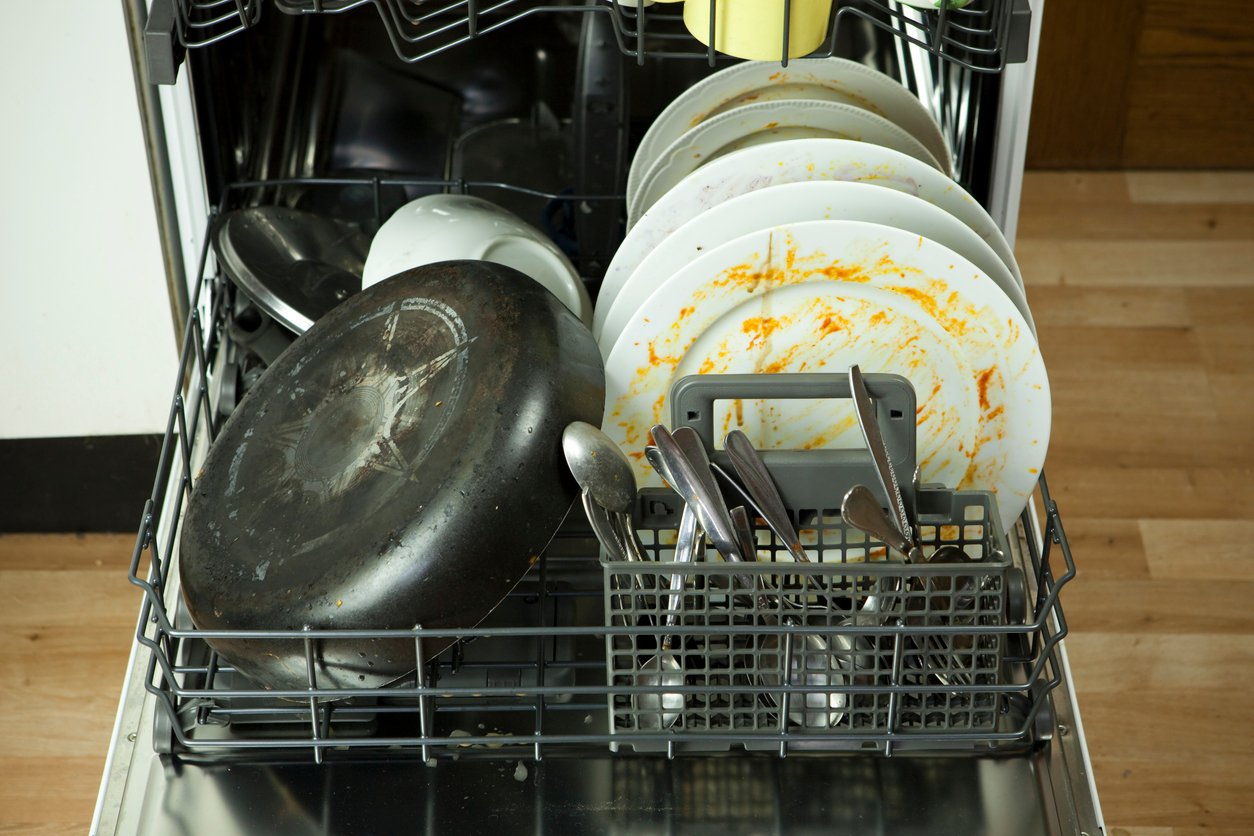 An open dishwasher filled with dirty plates covered in food residue, a frying pan, bowls, and assorted utensils in the cutlery basket, ready to be washed.