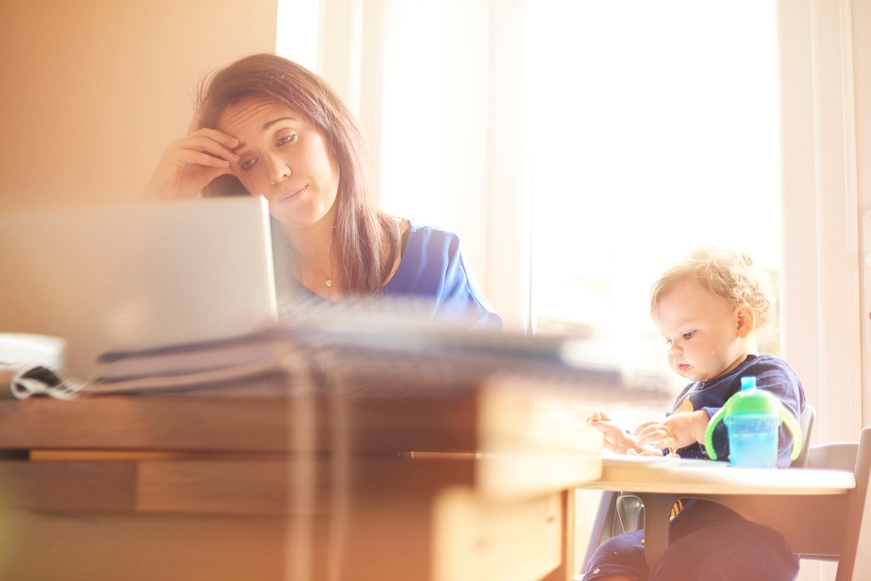 A woman sits at a desk with her hand on her forehead, looking tired while working on a laptop. A toddler sits next to her at the table, playing with a toy and a green bottle nearby. Sunlight streams through a window behind them.
