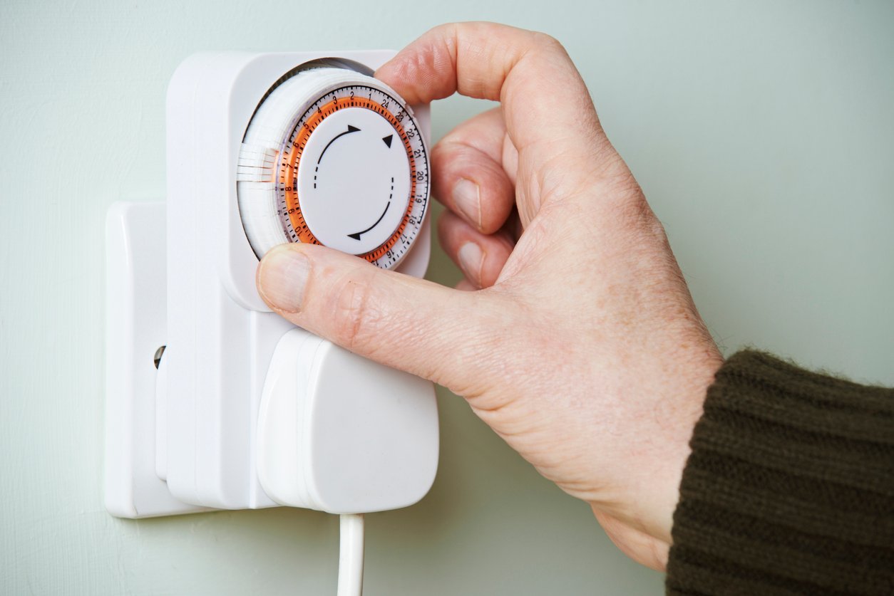 A hand adjusts a plug-in timer switch plugged into a wall socket, with a white dial displaying black numbers and orange markers.