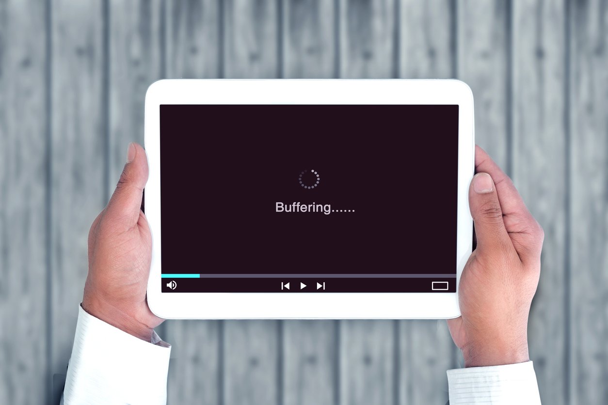 A person holds a tablet displaying a video player screen with the word "Buffering......" and a loading icon, against a wooden background.