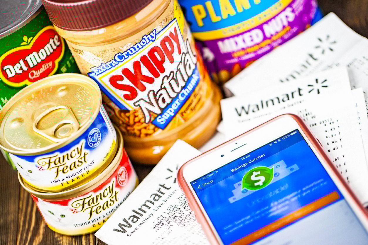 A close-up of canned food, peanut butter, and mixed nuts next to Walmart receipts. An iPhone displays a savings app. The products include Del Monte vegetables, Fancy Feast cat food, and Skippy Natural peanut butter.