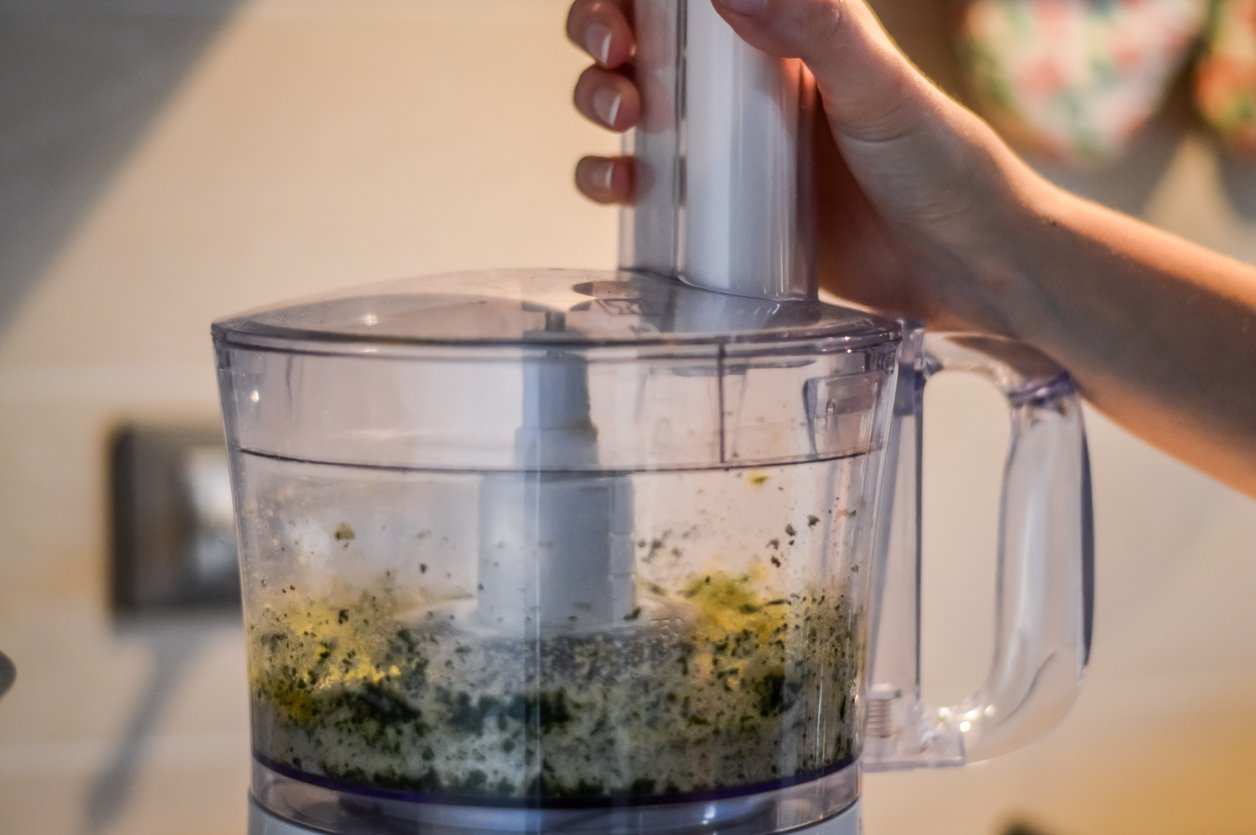 A hand holds the lid of a food processor blending green and white ingredients on a kitchen counter.