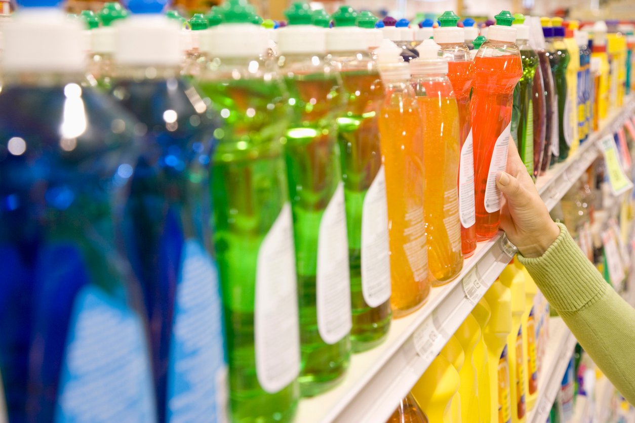 A person’s hand reaches for a bottle of colorful dish soap on a supermarket shelf lined with various cleaning products in different colors and shapes.