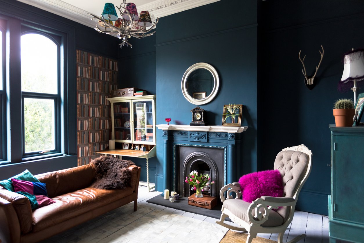 A stylish living room with dark blue walls, a brown leather sofa, an armchair with purple cushions, a decorative fireplace, a round mirror above it, a glass cabinet, bookshelf wallpaper, and a window letting in natural light.
