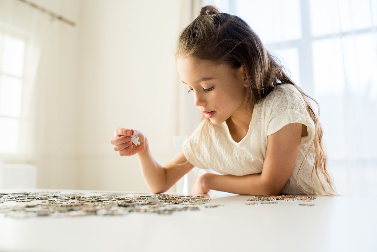 A young girl with long brown hair and a white shirt is concentrating as she assembles pieces of a jigsaw puzzle on a white table in a bright, sunlit room.