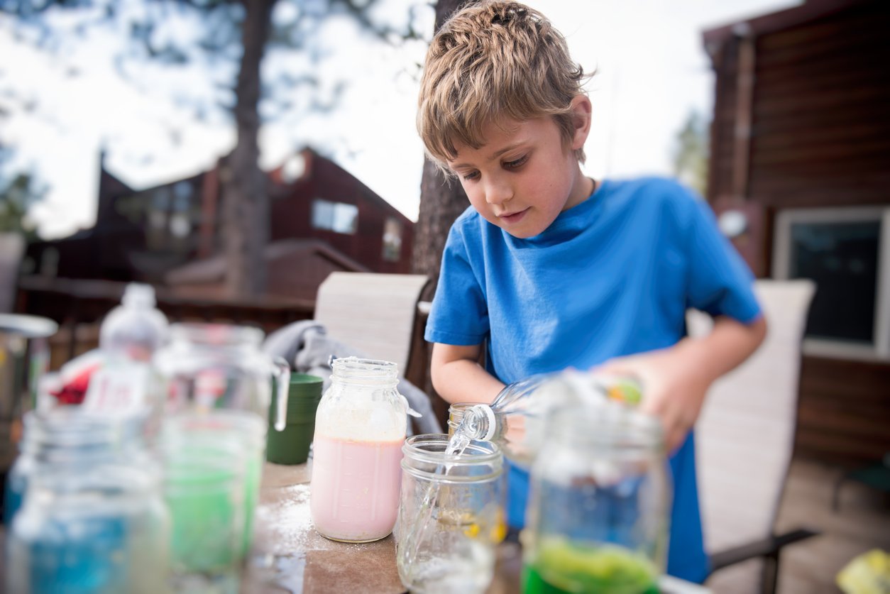 A young boy in a blue shirt pours liquid into a mason jar on an outdoor table filled with jars containing colorful drinks. Trees and wooden buildings can be seen in the blurred background.