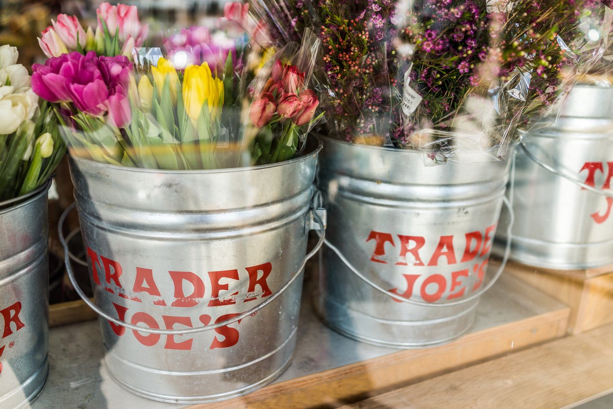 Three metal buckets labeled "Trader Joe's" are filled with colorful bouquets of flowers, including tulips and mixed blossoms, displayed on a wooden shelf.