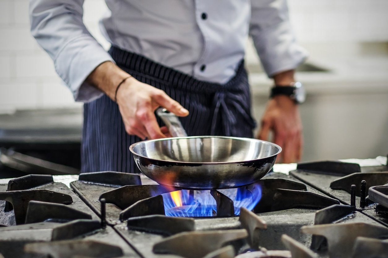 A chef in a white jacket and striped apron holds a metal frying pan over a blue gas flame on a stovetop in a commercial kitchen.