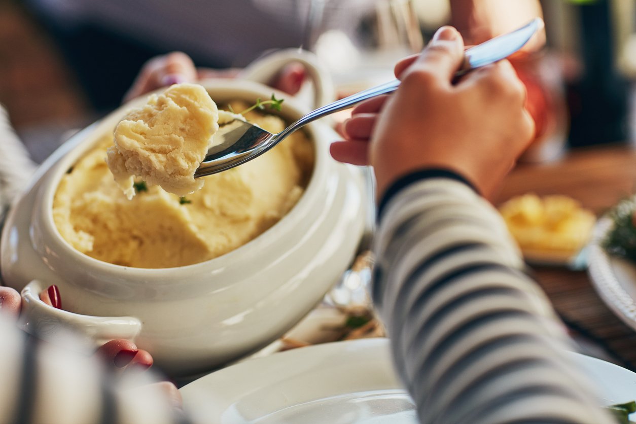 A person scoops creamy mashed potatoes from a large white bowl onto their plate. The scene suggests a shared meal, and the person is wearing a striped shirt.