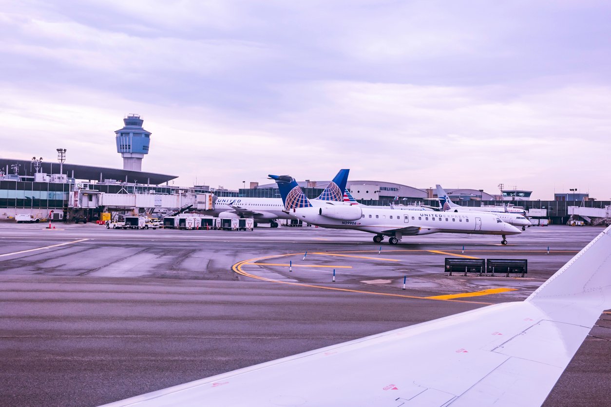 Two United Express airplanes are parked at airport gates near a terminal, with a control tower and ground service vehicles visible under a cloudy sky. The airplane wing is seen in the foreground.