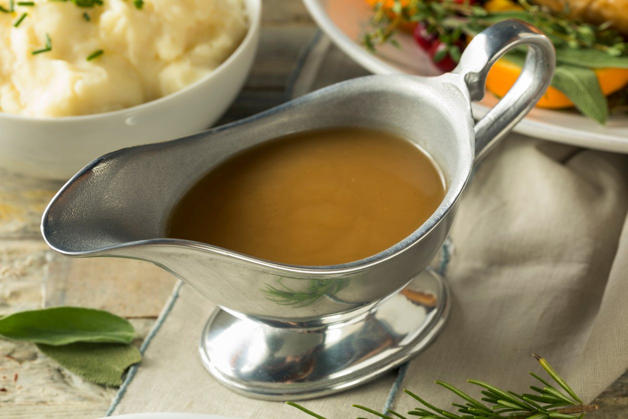 A silver gravy boat filled with brown gravy sits on a table, with a bowl of mashed potatoes and herbs visible in the background.