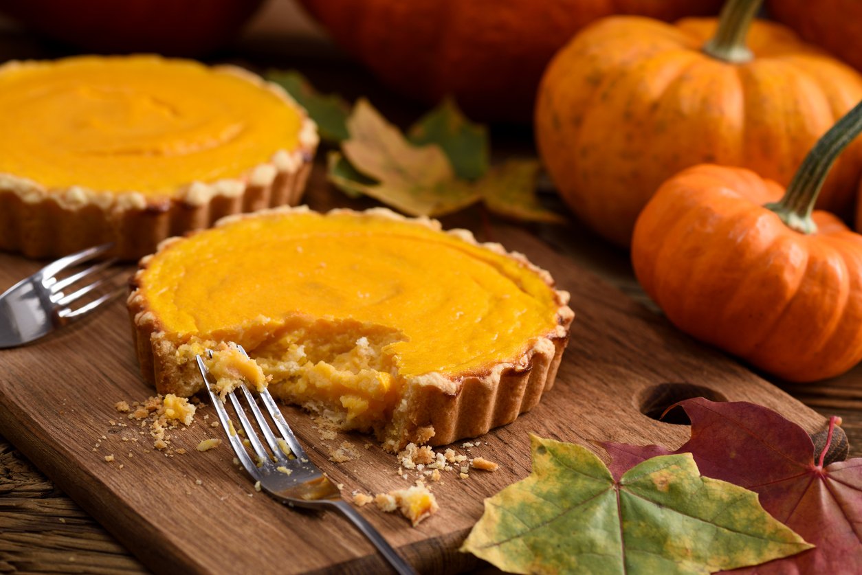Two mini pumpkin pies sit on a wooden board surrounded by autumn leaves and small pumpkins, capturing the cozy feel of Thanksgiving. With a bite taken out, the scene hints at memories made and moments shared—perfect for reflecting on Thanksgiving dinner cost.