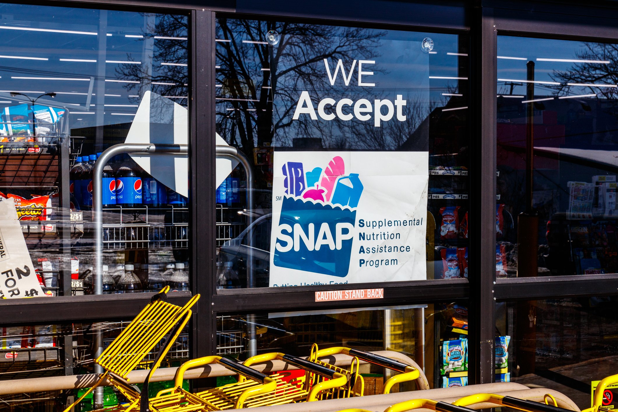 A store window displays a sign reading "We Accept SNAP Supplemental Nutrition Assistance Program". Shopping carts are visible outside the entrance, and store shelves with products can be seen inside.