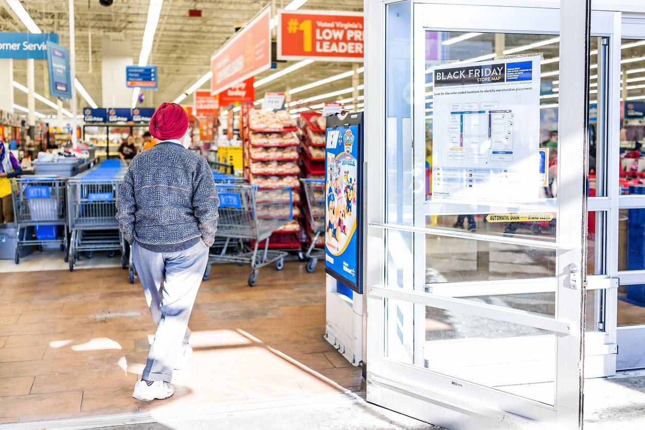 A person wearing a red hat and gray sweater walks into a brightly lit store with shopping carts and Black Friday sale signs visible near the entrance.