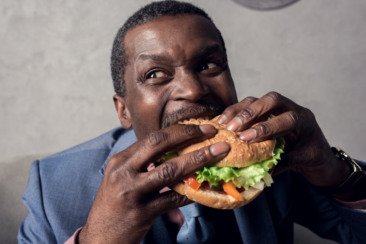 A man in a blue suit is excitedly taking a big bite of a large hamburger loaded with lettuce, tomato, and other toppings. He is holding the burger with both hands and looking up while eating.
