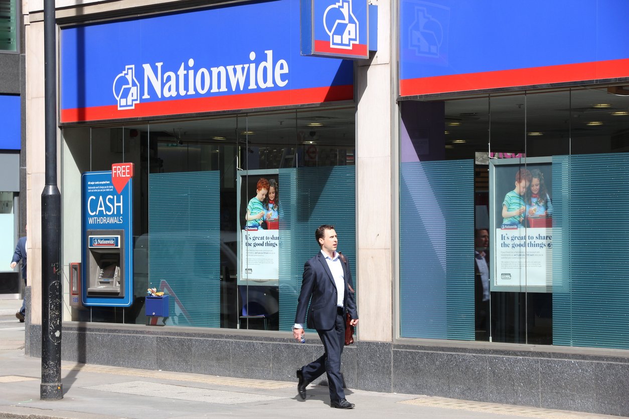 A man in a suit walks past a Nationwide bank branch with blue signage, large windows displaying posters, and an ATM labeled “Cash Withdrawals” on the exterior wall.
