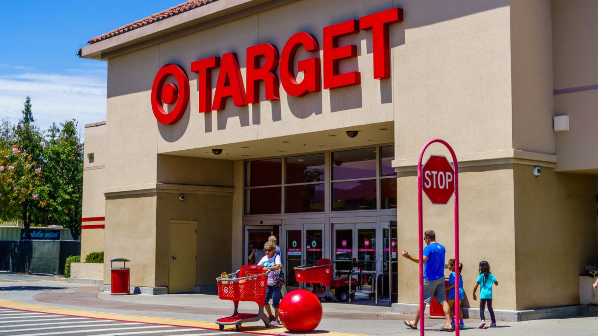 Customers with shopping carts are entering and exiting a Target store. The store has large red "TARGET" letters and logo above the entrance. A stop sign and large red decorative ball are in front of the building.