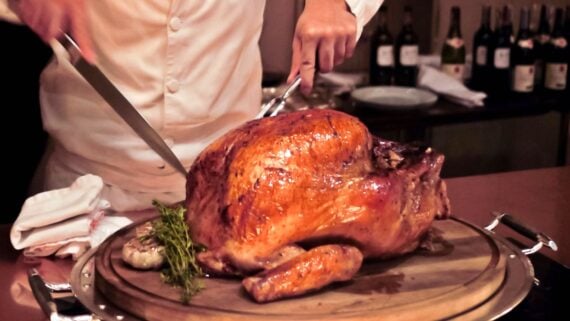 A chef in a white coat carves a golden-brown roasted turkey on a wooden board, with herbs and garlic nearby. Bottles of wine are visible in the blurred background.