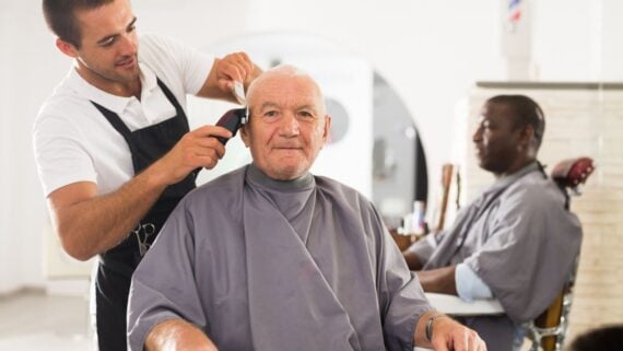 A barber in a white shirt trims the hair of an older man with clippers, while another man sits in the background, also wearing a cape, in a bright barbershop.