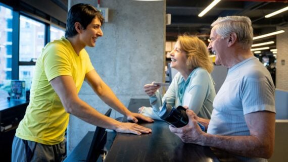 A young man in a yellow shirt smiles and talks with an older man and woman at a gym reception desk. The older couple appear happy and engaged in conversation. The man holds a water bottle.