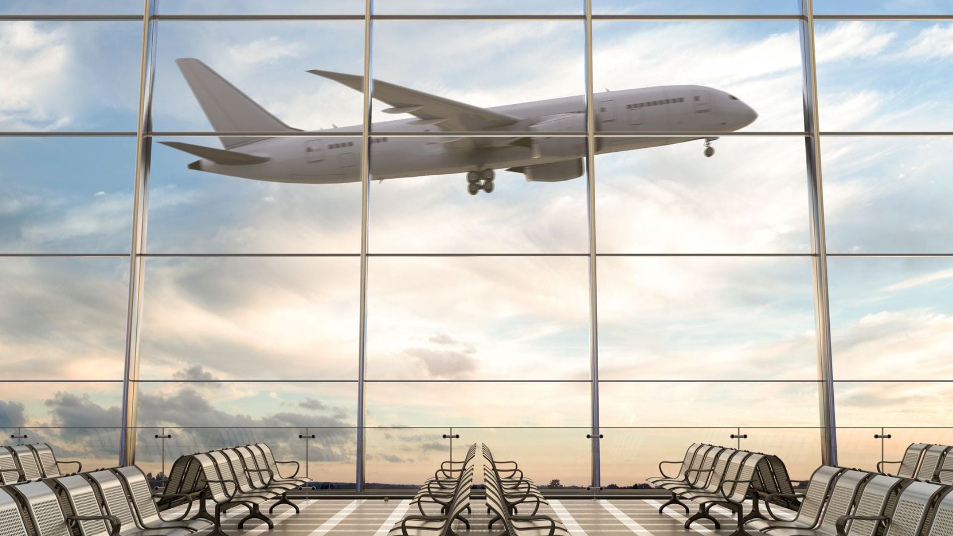 A large airplane flies past the windows of an empty airport terminal with rows of metal seats, under a sky with clouds and soft sunlight.