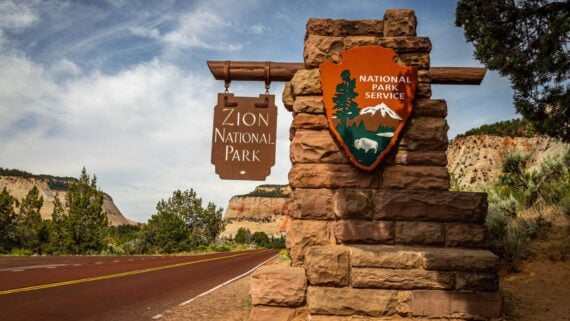 Stone entrance sign for Zion National Park with the National Park Service emblem, next to a red paved road, surrounded by trees, bushes, and rocky cliffs under a partly cloudy sky.