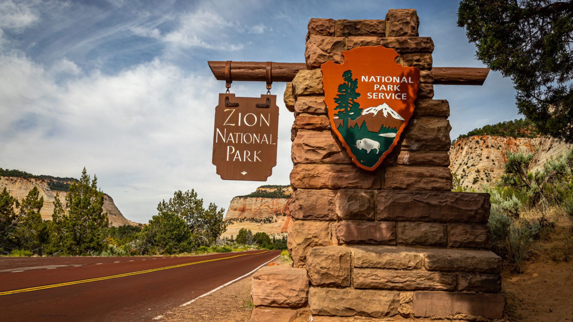 Stone entrance sign for Zion National Park with the National Park Service emblem, next to a red paved road, surrounded by trees, bushes, and rocky cliffs under a partly cloudy sky.