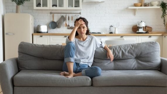 A woman sits barefoot on a gray couch in a modern kitchen, resting her elbow on her knee and her hand on her forehead, looking pensive or stressed. The kitchen has white cabinets and wooden countertops.