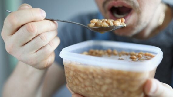 A person holding a plastic container of baked beans and eating them with a spoon, with their mouth open, ready to take a bite.