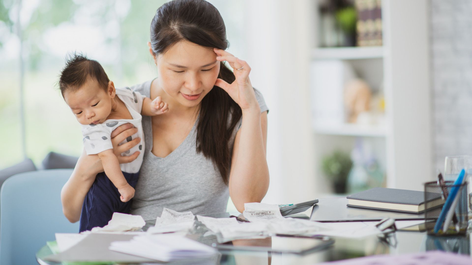 A woman holding a baby looks stressed while reviewing bills and paperwork at a table, with notebooks and papers scattered in front of her.