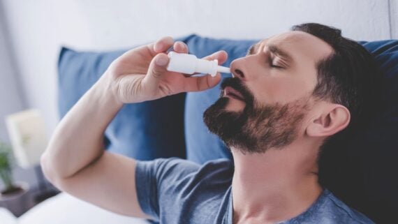 A man with a beard is sitting on a bed while using a nasal spray, tilting his head slightly back with his eyes closed.