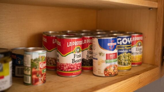 Several cans of food, including pink beans, black beans, green pigeon peas, and beef broth, are arranged on a wooden pantry shelf. The cans are various brands and sizes.