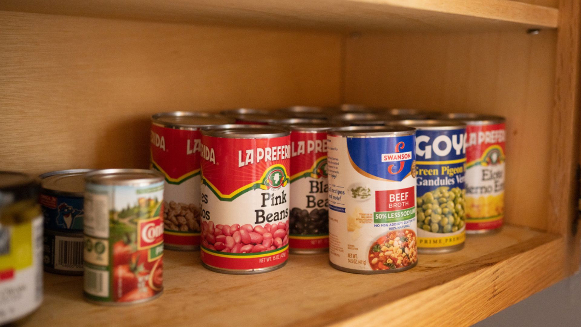 Several cans of food, including pink beans, black beans, green pigeon peas, and beef broth, are arranged on a wooden pantry shelf. The cans are various brands and sizes.