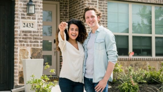 A smiling couple stands in front of a brick house, with one person holding up a key, suggesting they have just bought a new home. The house number 2432 is visible next to the door.