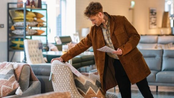 A man in a brown jacket examines the fabric of an armchair in a furniture store while holding a brochure. Sofas, pillows, and blankets are displayed around him.