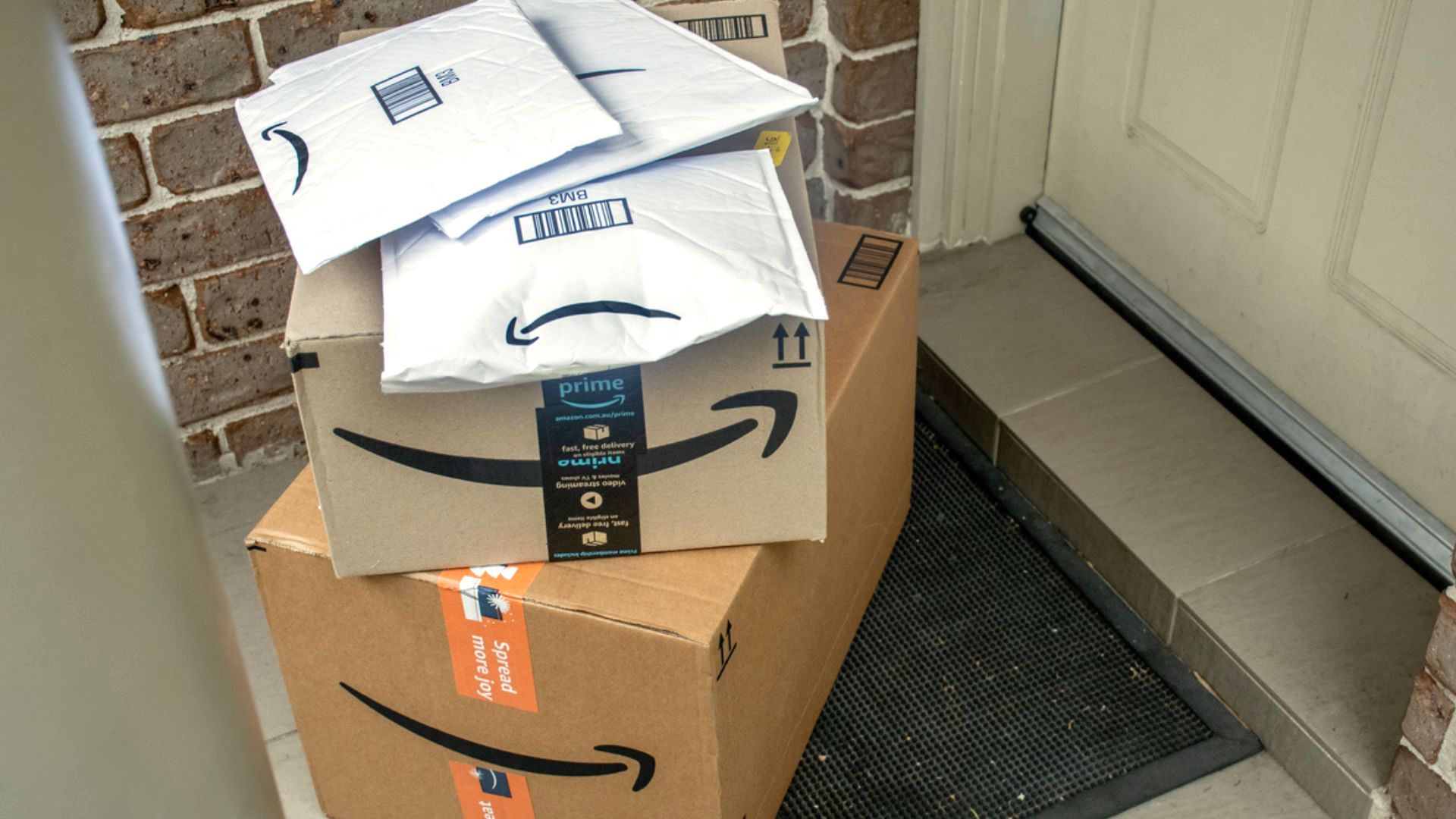 Several Amazon packages, including boxes and padded envelopes, are stacked on a black doormat in front of a white door with a brick wall background.