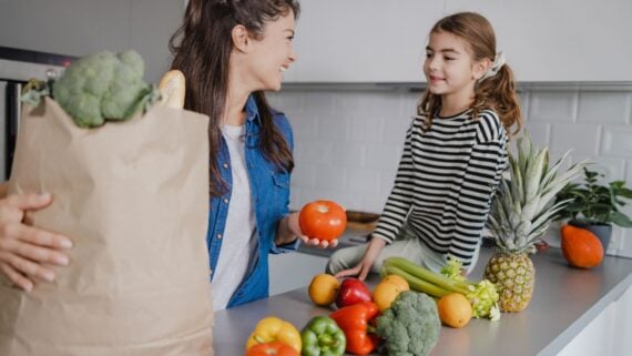 A woman and a young girl smile at each other in a kitchen. The woman holds a grocery bag and a tomato, while the counter is covered with fresh fruits and vegetables, including a pineapple, peppers, broccoli, and celery.