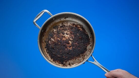 A hand holds a stainless steel frying pan with a heavily burnt and blackened bottom against a solid blue background.