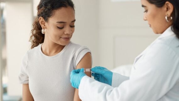 A healthcare professional wearing blue gloves applies a bandage to a woman's upper arm after vaccination. The woman is looking at her arm and smiling slightly, sitting in a bright, clinical setting.