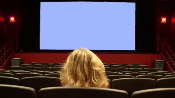 A person with blonde hair sits alone in a mostly empty movie theater, facing a large blank screen with rows of black seats around them.